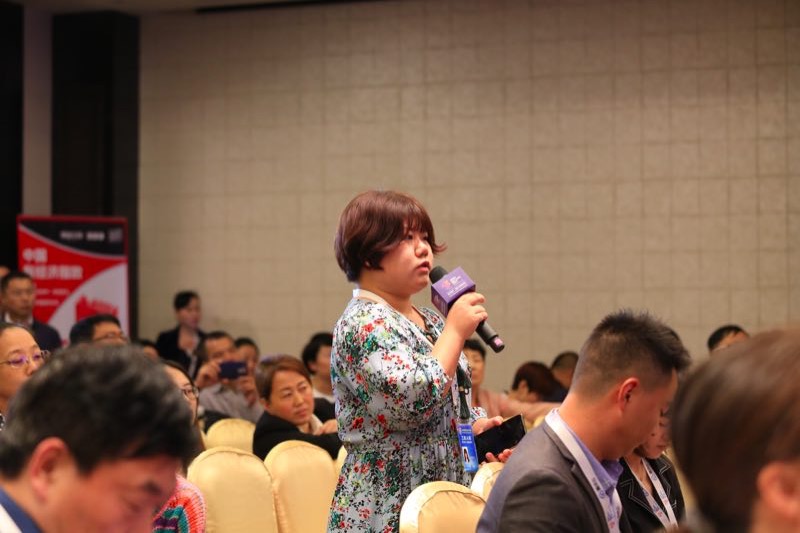 一个女人在房间里对着麦克风说话，背景中有人